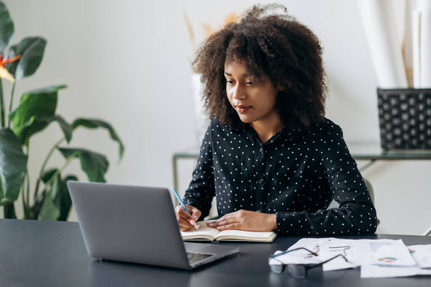 Young adult African American business woman in the office. Attractive confident smart woman top manager or trader, sits in front of a laptop, looks the screen, makes working notes in a notebook - Foto, immagini