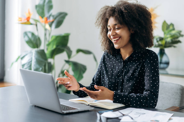 Succesvolle positieve jonge volwassen vrouw Afrikaanse vrouw freelancer, manager, CEO, zittend in het kantoor op laptop, praten op video gesprek met klant of medewerkers, bespreken van zakelijke strategie, gebaren, glimlach - Foto, afbeelding