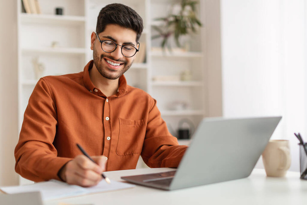 Sourire homme arabe dans des lunettes à l'aide d'un ordinateur portable et de prendre des notes - Photo, image