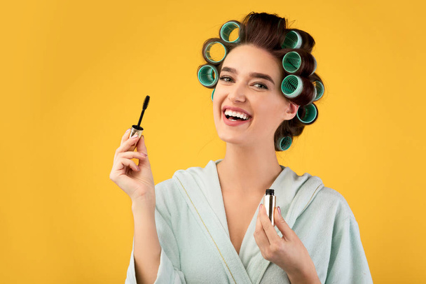 Happy Woman With Curlers Holding Mascara Over Yellow Studio Background - Foto, afbeelding