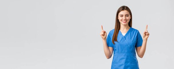 Gesundheitshelfer, Virusvorbeugung, Versicherungs- und Medizinkonzept. Attraktive Krankenschwester oder Ärztin im blauen Peeling, mit erhobenen Fingern lächelnd, Werbebanner, medizinische Pillen, grauer Hintergrund - Foto, Bild