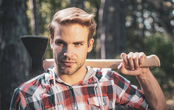 A handsome young man with a beard carries a tree. Handsome Woodworkers lumberjack plaid shirt holding the axe on green nature background. - Photo, image