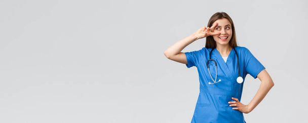 Медицинские работники, здравоохранение, ковид-19 и концепция вакцинации. Веселая и энергичная молодая женщина-врач, красивая медсестра в синем халате, стетоскоп, показать мир возле глаз и улыбаясь оптимистично
 - Фото, изображение