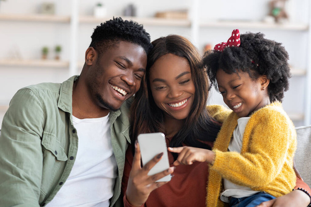 Tolle App. Glückliche schwarze Familie mit kleiner Tochter nutzt Smartphone zu Hause - Foto, Bild