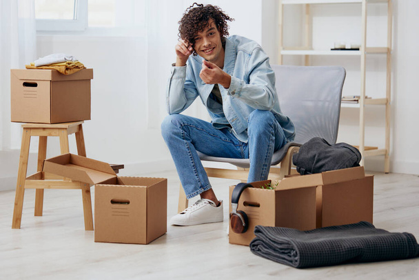 A young man sitting on a chair unpacking with box in hand moving interior - Photo, Image