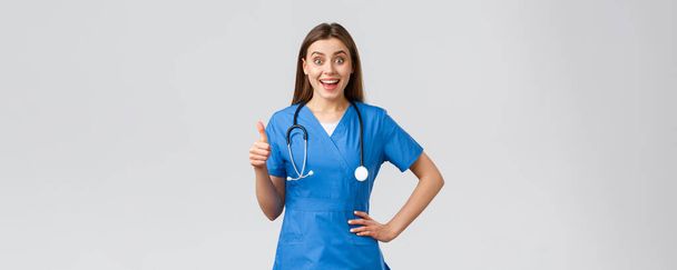 Trabajadores médicos, atención médica, covid-19 y concepto de vacunación. Enfermera emocionada sorprendida o médico en uniformes azules con estetoscopio, mostrar los pulgares hacia arriba en la aprobación, como idea, sonriendo optimista
 - Foto, Imagen