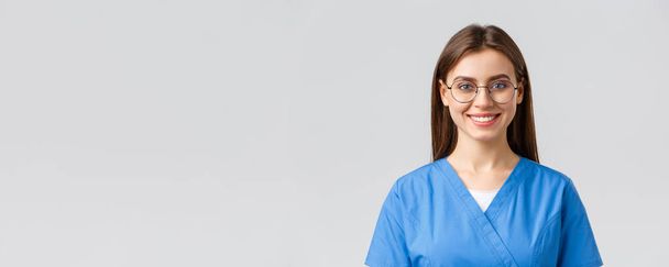医療従事者、医療、保険およびcovid-19パンデミックの概念。陽気な楽観的な女性看護師,医師やクリニックでのインターンは、スクラブや眼鏡をかけて,正のまま,アップビート笑顔 - 写真・画像