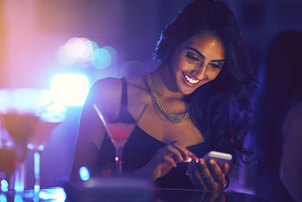 Kom hier, dit feest is geweldig. Shot van een vrouw met behulp van haar mobiele telefoon tijdens het zitten aan een tafel in een nachtclub. - Foto, afbeelding