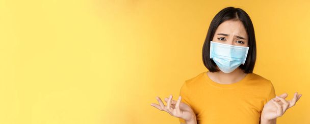 Close up portrait of confused asian woman in medical face mask, shrugging shoulders and looking puzzled, standing against yellow background - Fotó, kép