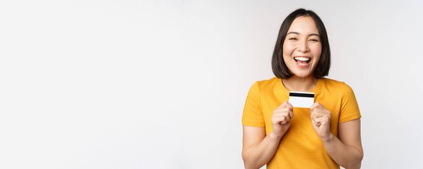 Portrait of beautiful korean girl holding credit card, recommending bank service, standing in yellow tshirt over white background - Foto, Imagen