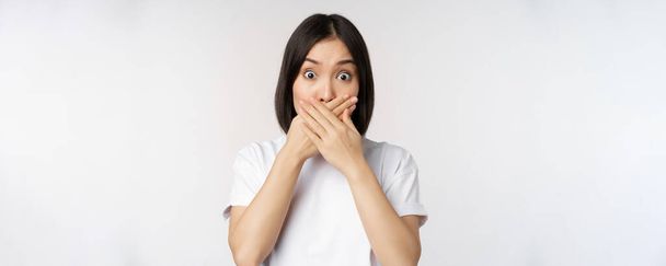 Bild einer schockierten jungen asiatischen Frau, die den Mund zuhält, erschrocken, sprachlos schaut und vor weißem Hintergrund steht - Foto, Bild
