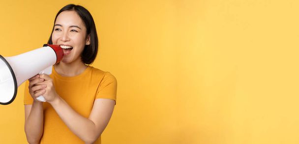 ¡Anuncio! Mujer asiática feliz gritando fuerte en megáfono, reclutando, protestando con el altavoz en las manos, de pie sobre el fondo amarillo - Foto, imagen