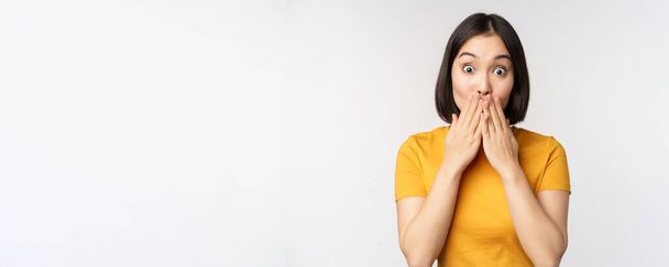 Überraschte asiatische Frau keuchend, Mund mit Händen bedeckt und erstaunt in die Kamera blickend, in gelbem T-Shirt, vor weißem Hintergrund stehend - Foto, Bild