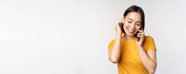 Romantikus és aranyos ázsiai lány veszi fel a telefont, beszél a mobil, mosolygós kacér, álló sárga póló ellen fehér háttér - Fotó, kép
