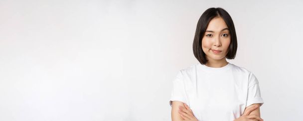 Close up of korean girl, looking skeptical, cross arms on chest and smirk, stare with disbelief at camera, standing over white background - Photo, Image