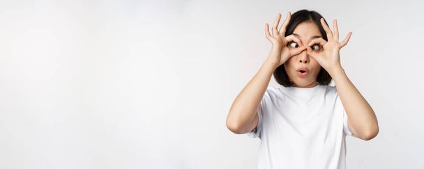 Αστεία νεαρή Ασιάτισσα, Κορεάτισσα που κάνει χειρονομίες γυαλιών, που δείχνει χαρούμενη στην κάμερα, στέκεται πάνω από λευκό φόντο - Φωτογραφία, εικόνα