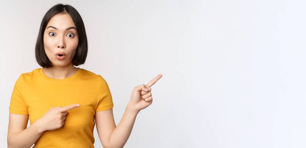 Amazed young asian woman, showing advertisement aside, pointing fingers right at promotion text, brand logo, standing happy against white background - Photo, Image