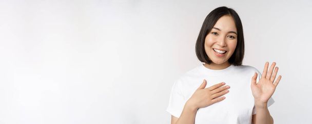 Portret van een prachtig Koreaans meisje dat haar hand opsteekt, zich voorstelt, haar hand op het hart legt, begroet, over een witte achtergrond staat - Foto, afbeelding