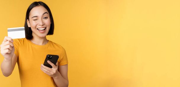 Chica asiática alegre sonriendo, mostrando la tarjeta de crédito y el teléfono inteligente, recomendando la banca del teléfono móvil, de pie sobre el fondo amarillo - Foto, Imagen
