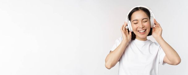 Modernes asiatisches Mädchen tanzt, hört Musik mit Kopfhörern, lächelt glücklich, steht im T-Shirt vor weißem Hintergrund - Foto, Bild