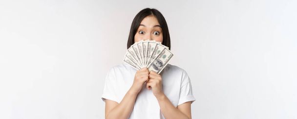 Щаслива азіатська дівчина тримає гроші, готівка біля обличчя, концепція мікрокредитування та фінансів, білий студійний фон
 - Фото, зображення