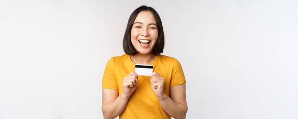 Portrait of beautiful korean girl holding credit card, recommending bank service, standing in yellow tshirt over white background - Photo, Image