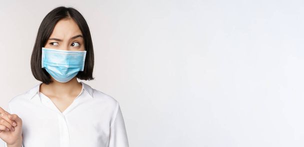 Imagen de una joven asiática con máscara facial médica, apuntando a la izquierda y mirando con sospechosa expresión confusa, de pie sobre fondo blanco - Foto, imagen