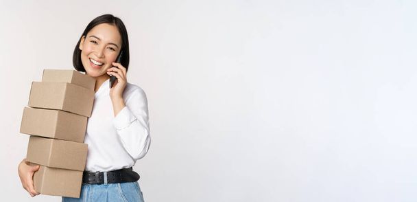 Imagen de joven mujer de negocios asiática contesta llamada telefónica mientras lleva cajas para la entrega, posando sobre fondo blanco - Foto, Imagen