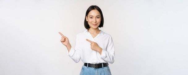 Bild einer lächelnden jungen Bürokauffrau, asiatischer Unternehmer zeigt mit dem Finger nach links, zeigt Kundeninformationen, Banner seitlich auf Kopierfläche, weißer Hintergrund - Foto, Bild
