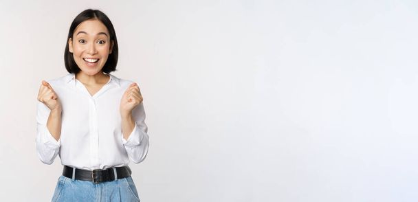 Enthusiastic asian woman rejoicing, say yes, looking happy and celebrating victory, champion dance, fist pump gesture, standing over white background - Fotó, kép