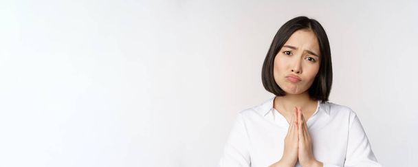 primer plano retrato de asiático mujer mendigando, pidiendo smth, decir por favor con tímido triste cara, necesidad de ayuda, de pie sobre blanco fondo - Foto, Imagen