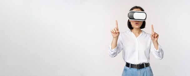 Bild einer asiatischen Frau in vr Brille, die mit dem Finger nach oben zeigt, erstaunt und überrascht guckt. Mädchen mit Virtual-Reality-Headset, Konzept der Zukunft und Kommunikation, weißer Hintergrund - Foto, Bild