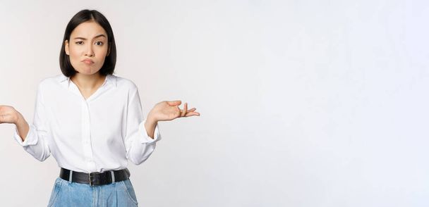 Image of confused asian office manager, office lady shrugging shoulders and looking clueless, standing puzzled against white background - Foto, Imagen