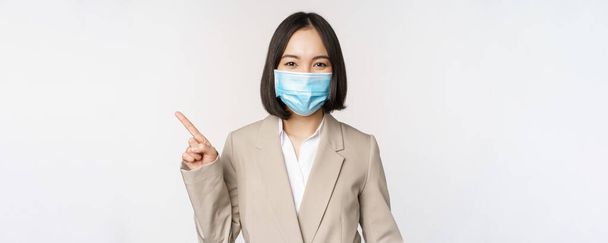 Coronavirus and work concept. Portrait of woman in medical face mask, pointing finger left, showing logo or banner, advertisement, white background - Photo, Image