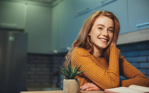 Smart redheaded smiling student sitting at work table looking at camera. Fashion model. Pretty young woman. Beautiful female person. - Foto, afbeelding