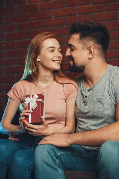 Terwijl het vieren van Saint Valentines Day man geeft een verrassing cadeau aan de vrouw thuis zitten op de bank en kijken met liefde naar elkaar. - Foto, afbeelding