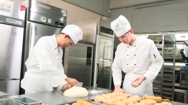 Zwei professionelle asiatische Köche in weißen Kochuniformen und Schürzen kneten Teig und Eier, bereiten Brot und frische Backwaren zu, backen im Ofen in der Edelstahlküche des Restaurants. - Filmmaterial, Video