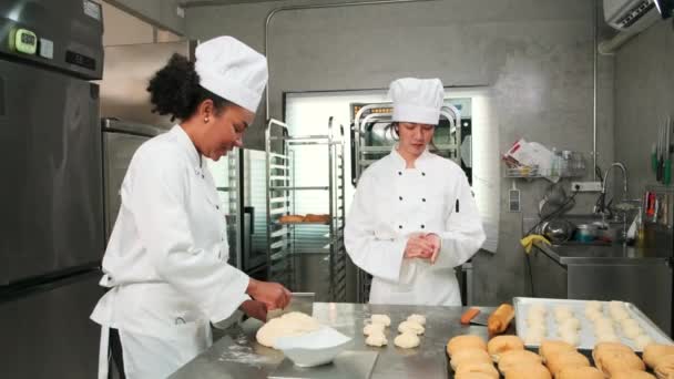 Zwei professionelle Köchinnen in weißen Kochuniformen und Schürzen kneten Teig und Eier, bereiten Brot, Plätzchen und frische Backwaren zu und backen im Ofen in der Edelstahlküche eines Restaurants. - Filmmaterial, Video