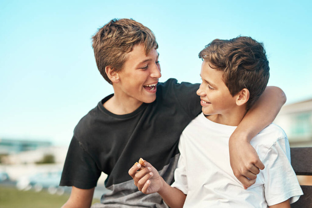 Hey, want to hear a joke. Shot of two happy brothers sitting on a bench by the beach together. - Photo, Image
