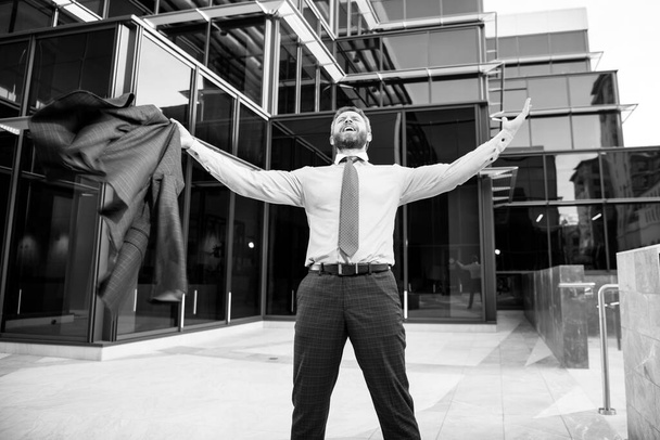 Excited businessman keeping arms raised and expressing positivity while standing outdoors with office building. - Foto, Imagen
