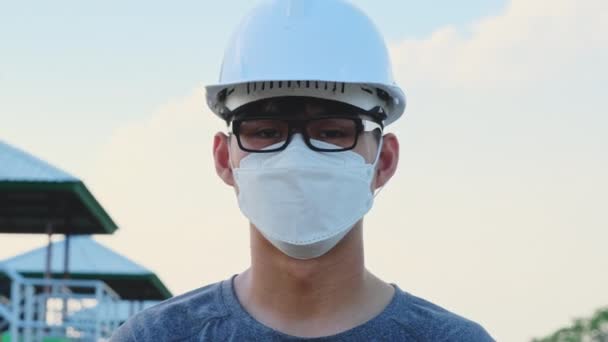 Jonge Aziatische ingenieur met een helm en masker kijkt en glimlacht naar de camera op de achtergrond van de dam. Een jonge ingenieur werkt in een dam tijdens de coronavirus uitbraak. - Video