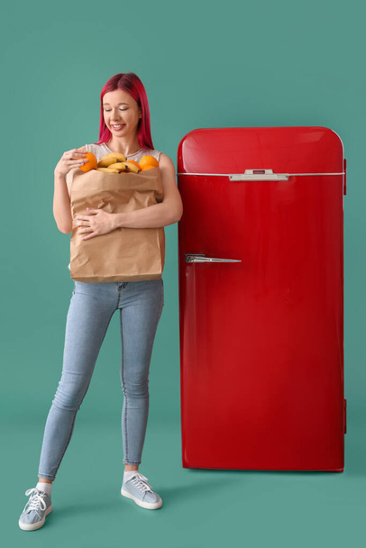 Όμορφη νεαρή γυναίκα με προϊόντα κοντά στο κόκκινο ψυγείο σε πράσινο φόντο - Φωτογραφία, εικόνα