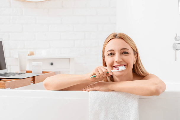 Beautiful mature woman brushing teeth while taking bath in morning - Photo, Image