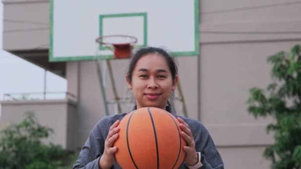 Mujer asiática sosteniendo baloncesto mirando a la cámara en el patio de baloncesto al aire libre. - Metraje, vídeo