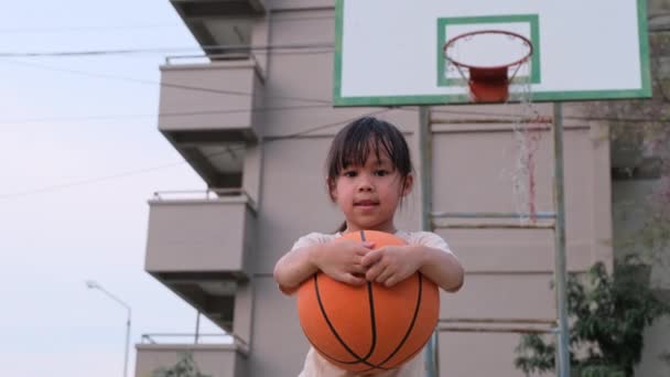 Веселая милая девушка, держащая баскетбол, смотрит в камеру на открытой баскетбольной площадке. - Кадры, видео