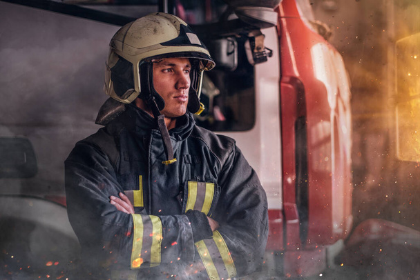 Feuerwehrmann in Schutzuniform steht neben einem Feuerwehrfahrzeug in der Garage einer Feuerwehr, verschränkt die Arme und blickt zur Seite - Foto, Bild
