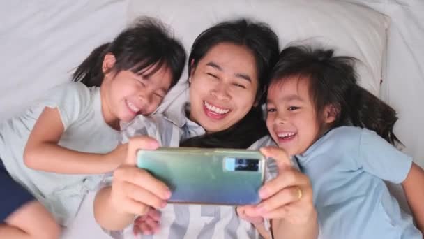 Feliz familia asiática disfrutando con smartphone en casa acogedora. Madre sonriente e hijas lindas usando el teléfono, Tome un selfie o video clip para recordar en la cama en casa. Buen momento en casa. - Metraje, vídeo