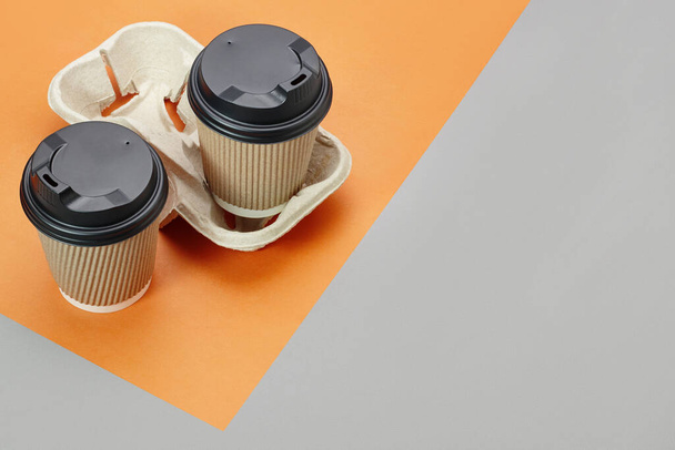 Tasses à café jetables en papier avec capuchons noirs sur support en carton sur fond orange et gris, endroit vide pour le texte. Café à emporter. - Photo, image