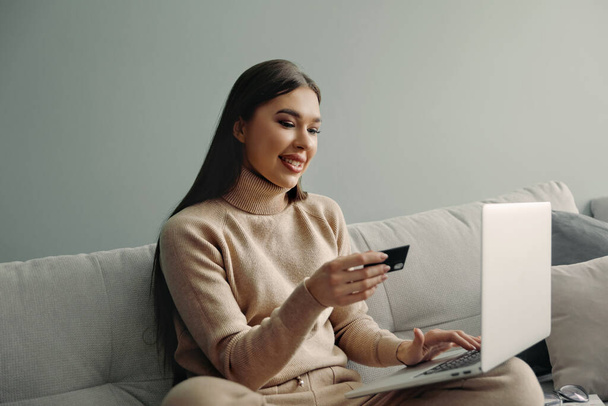 Woman is holding credit card and using laptop computer  sitting on couch at home,. Online shopping concept.  - Фото, зображення