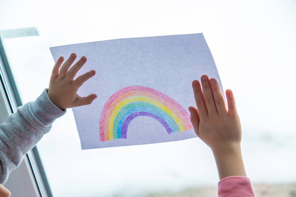 ζωγραφισμένο ουράνιο τόξο σε ένα φύλλο. Τα παιδιά κρατούν ένα φύλλο με ένα ουράνιο τόξο κοντά στο παράθυρο. επιλεκτική εστίαση - Φωτογραφία, εικόνα